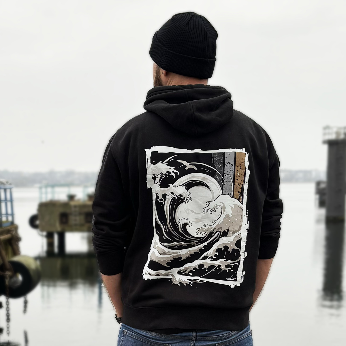 Big-Wave-Wellenmotiv auf schwarzem Kapuzenoberteil – Rückenansicht am Model im Hafen